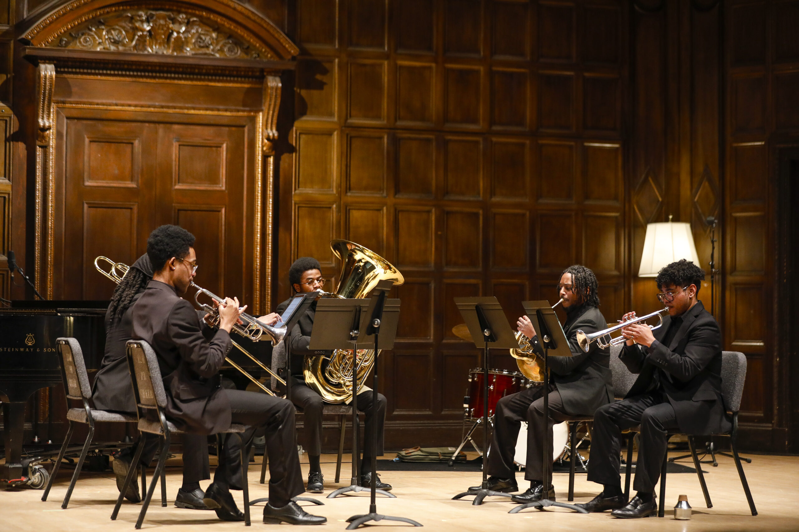 A group of five students on stage in Kilbourn Hall at Eastman School of music. They are seated in a semi-circle with music stands in front of them and are playing trumpet, trombone, tuba, French horn, and trumpet respectively from left to right.
