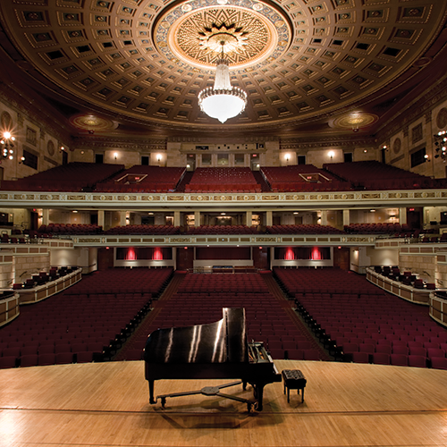 A photo looking out from the stage of and empty Kodak hall. A piano stands alone on the stage in the center of the image.