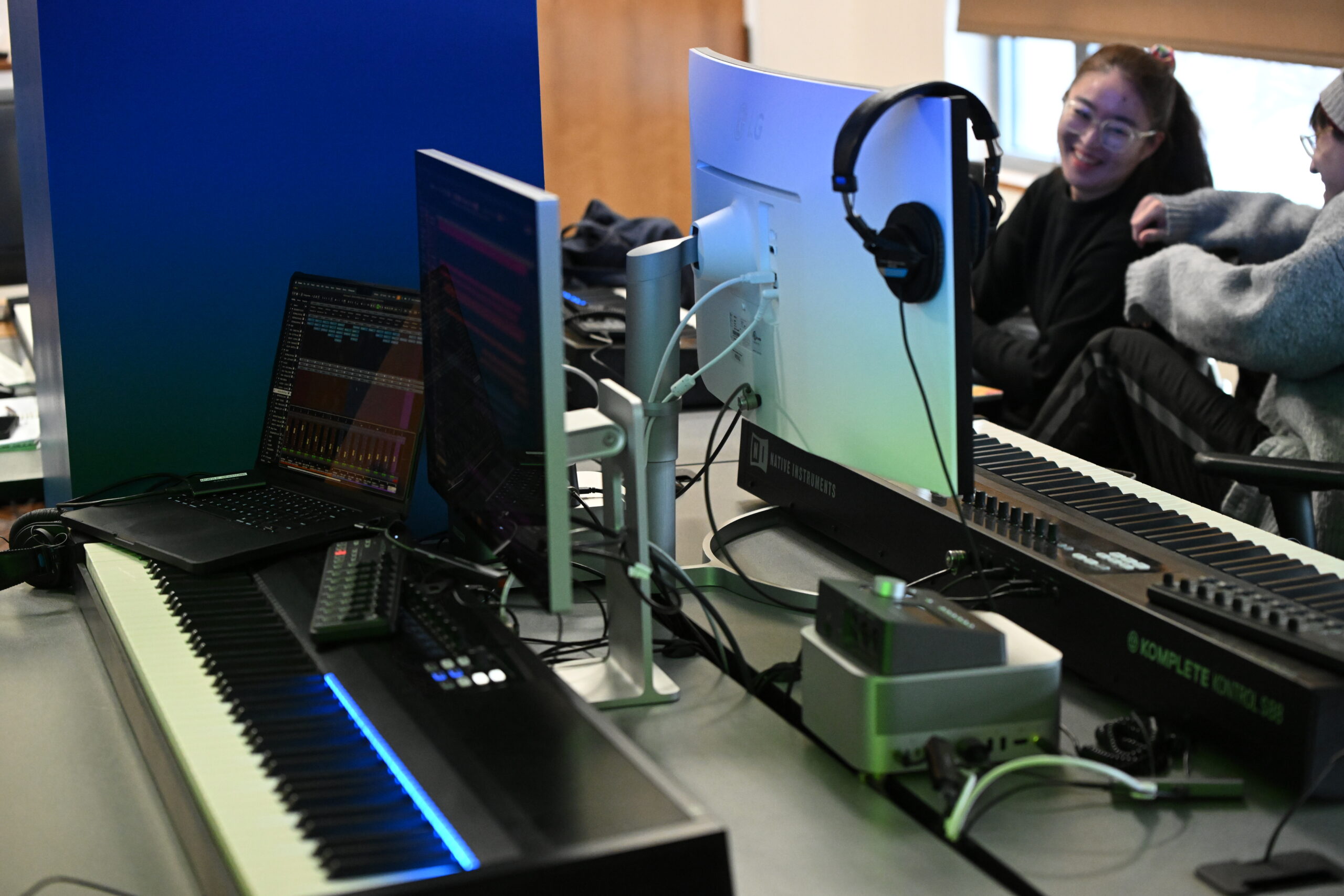 A media lab with two midi keyboard and monitor setups, a laptop with its screen facing the camera shows a program open with digital audio sliders on the screen. Two students are out of focus and laughing in the background