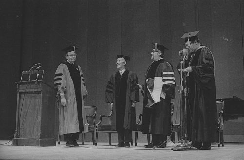 Conferral of an honorary doctoral degree on Shinichi Suzuki. Professor Donald Shetler hoods the newly named Dr. Suzuki; University President Robert Sproull stands at far left, and ESM Director Walter Hendl stands at far right.