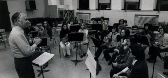 Verne Reynolds, standing, smiles while rehearses a choir of 15 horn students in a classroom at the Eastman School of Music.