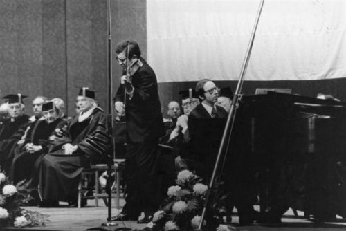 Zvi Zeitlin and Barry Snyder perform during a convocation in honor of Her Excellency Golda Meir, Eastman Theatre, December 16, 1974. Photograph from ESPA 37-13 (8x10). Zvi Zeitlin performs on violin with Barry Snyder at the piano during a convocation ceremony in Eastman Theatre, with honoree Golda Meir and faculty wearing regalia in the background.