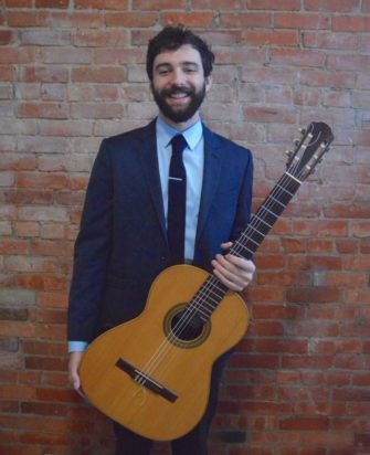 Man standing in front of a brick wall holding guitar and smiling.