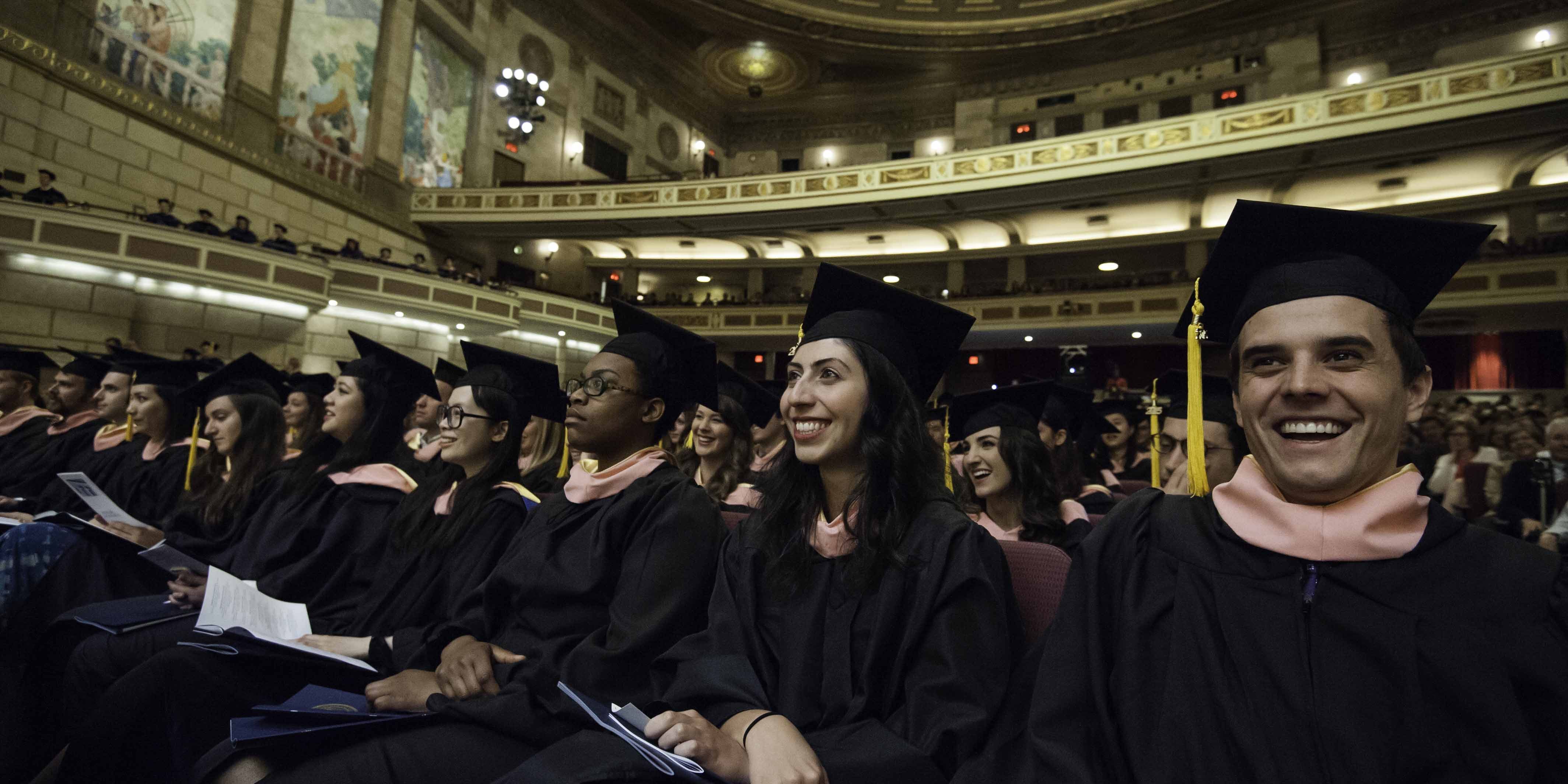 Commencement 2015 students Listening A row of smiling students in graduation caps and gowns in the theater.