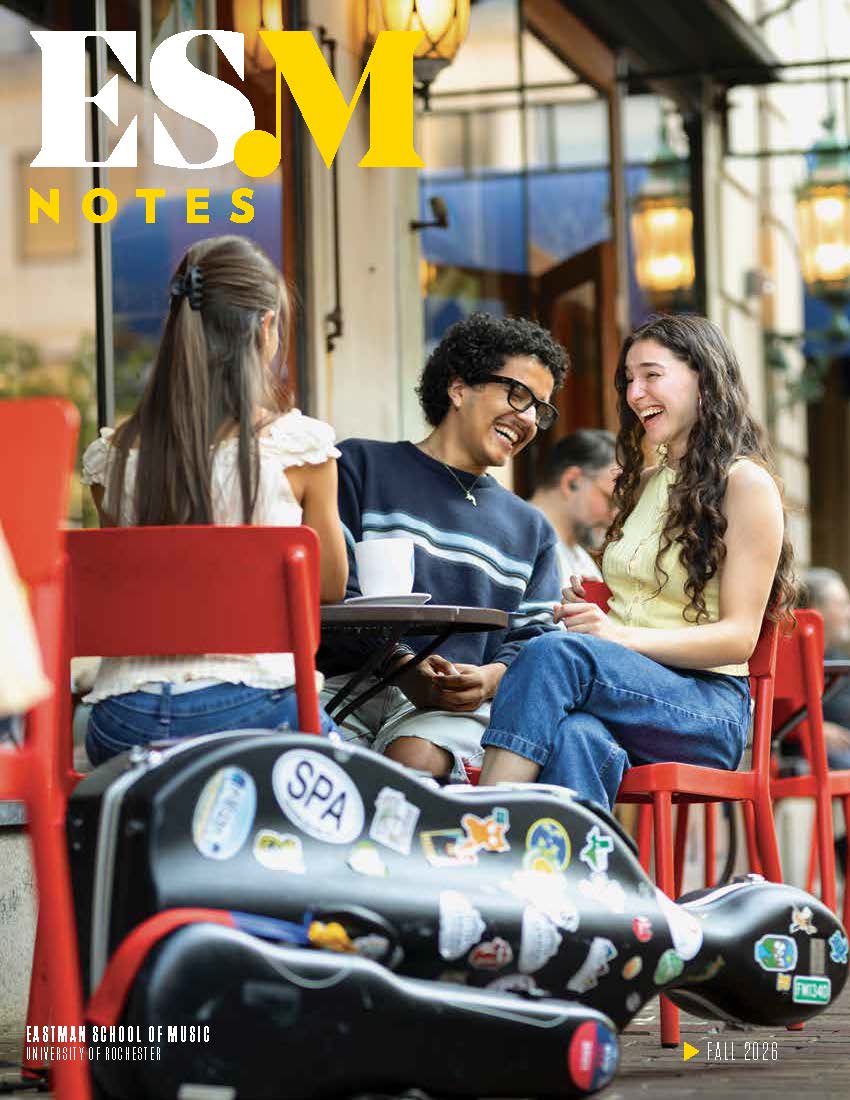 The Cover of Eastman NOTES Spring 2026 issue. The cover image depicts students sitting and chatting outside of Eastman School of Music on a bright spring day with an instrument case sitting in the foreground next to them
