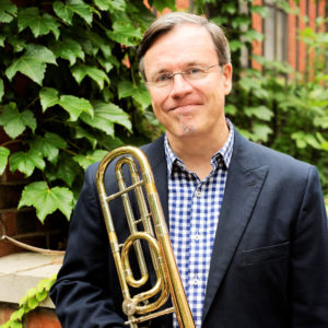 A photograph of Mark Kellogg holding a trombone in front of an Ivy covered building. Photo by Gerry Szymanski