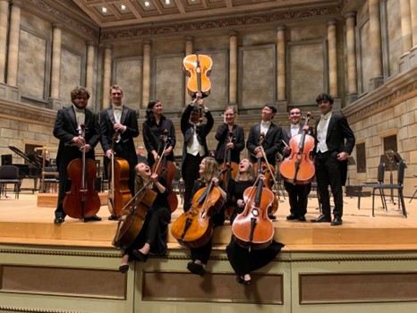 Cellists dressed in black concert attire on stage. One cello is held up in the air.