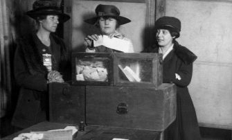A vintage photo of three Suffragettes around a voting box.