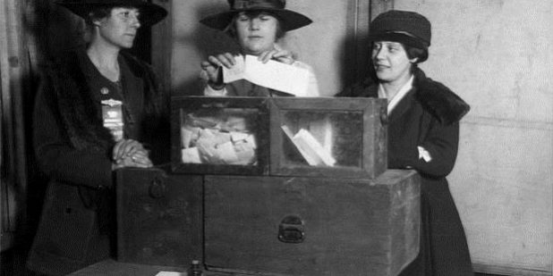 A vintage photo of three Suffragettes around a voting box.