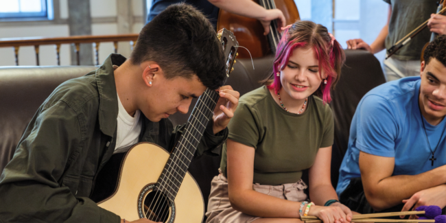 three students sit on a couch (two male, one female), one male student plays guitar while the others watch.