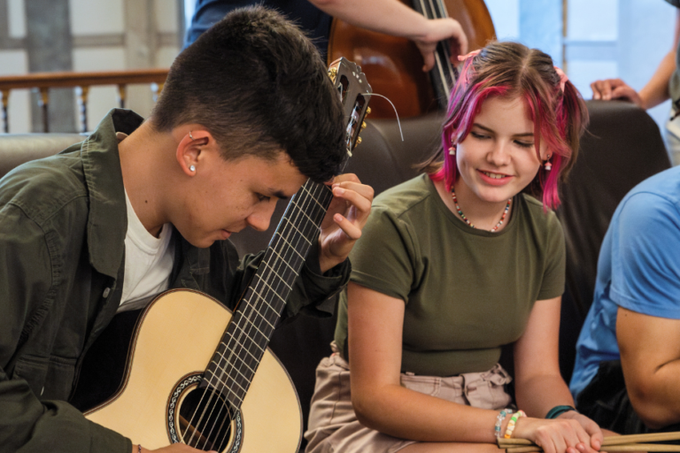 three students sit on a couch (two male, one female), one male student plays guitar while the others watch.
