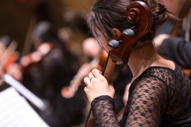 A woman in a black dress playing cello in an orchestra setting.