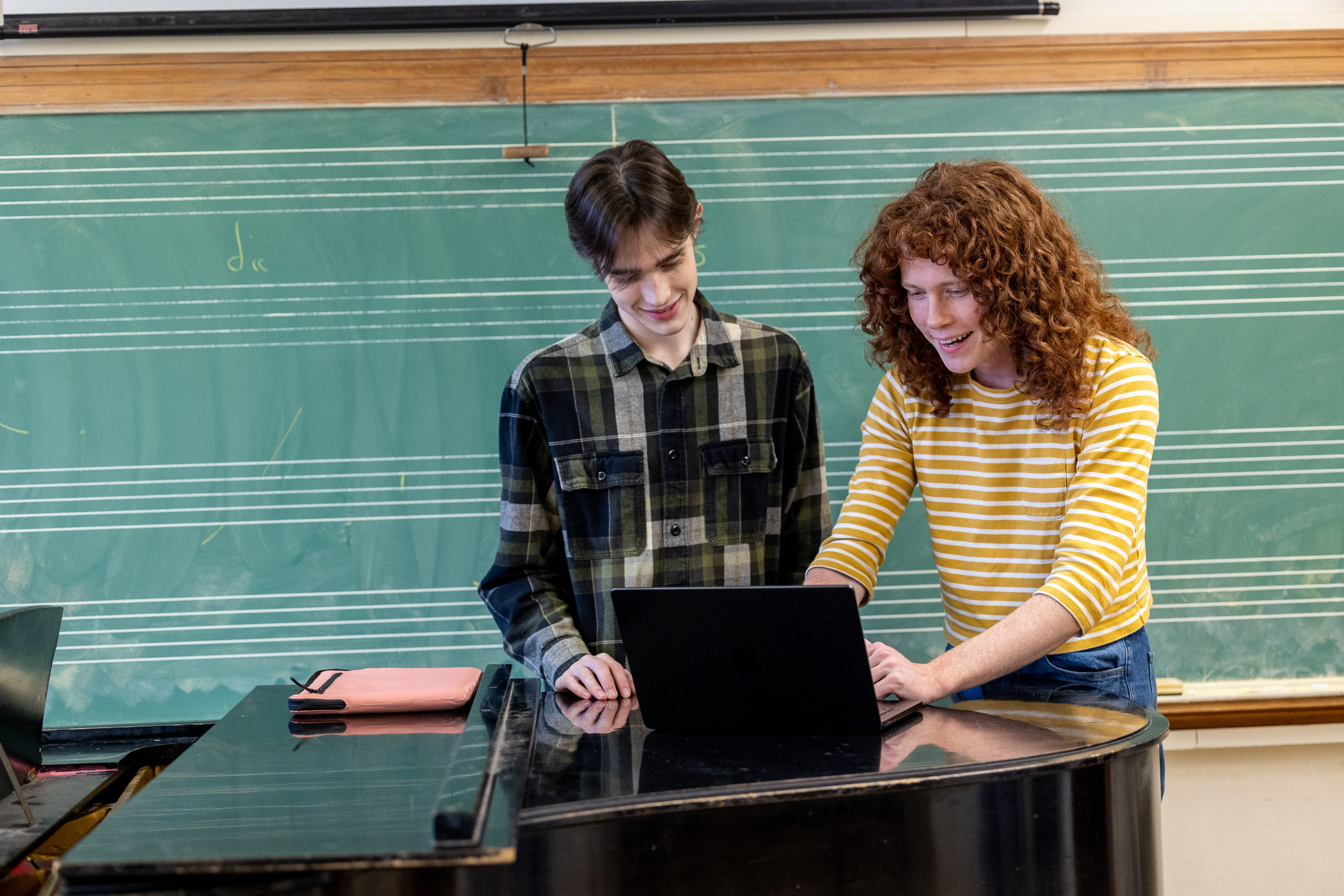 Two students at laptop, leaning on piano, in front of chalkboard. Student on the right has long, red curly hair; student on the left has short, black hair.